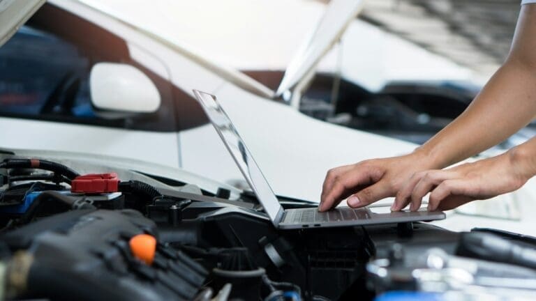 Car mechanic using laptop computer examining tuning fixing repairing car engine in garage.