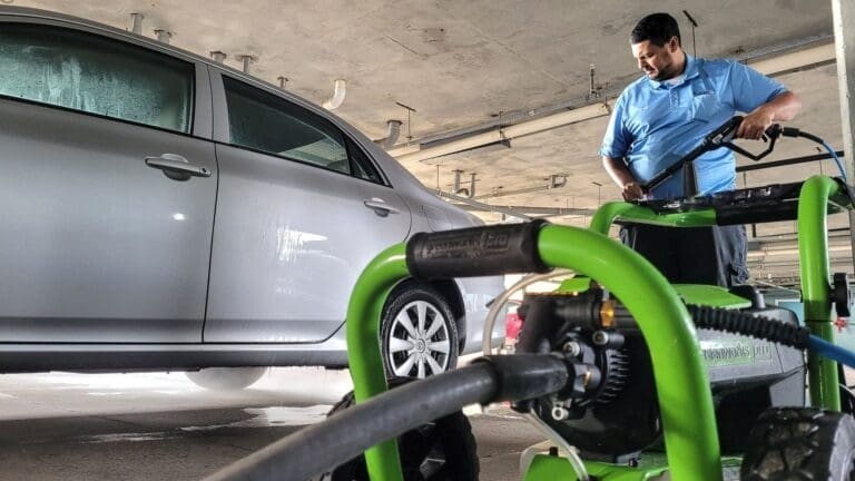 Millennial maintenance man at condo uses digital power washer to clean a Toyota corolla in garage.